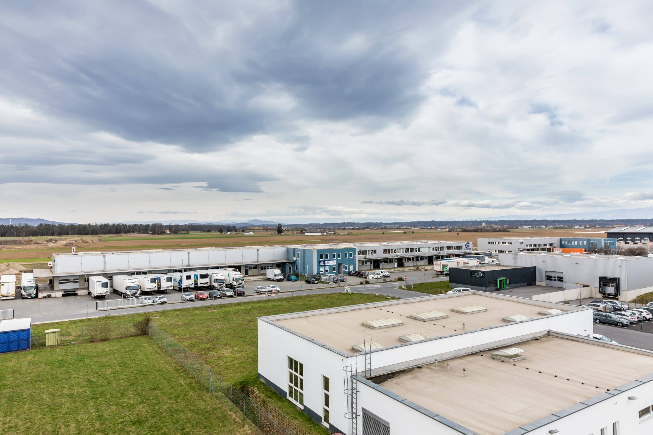 Industriegebiet mit Lagerhallen, LKWs und Parkplatz unter bewölktem Himmel.