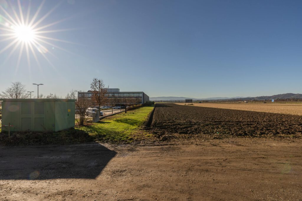 Großes, leeres Feld an sonnigem Tag, mit angrenzender Straße und entfernten Bergen im Hintergrund.