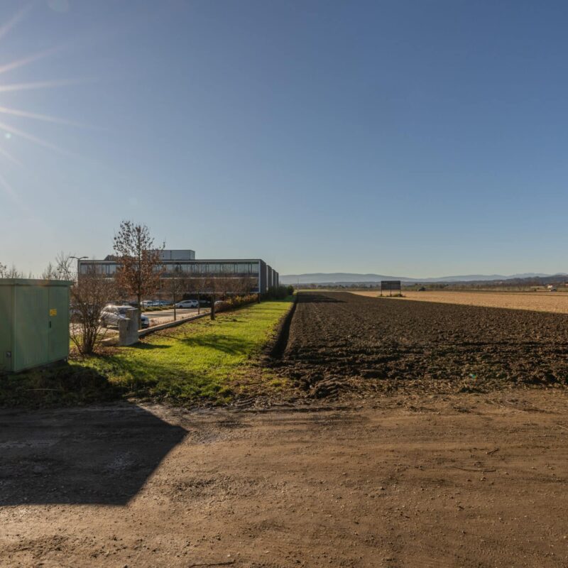Großes, leeres Feld an sonnigem Tag, mit angrenzender Straße und entfernten Bergen im Hintergrund.