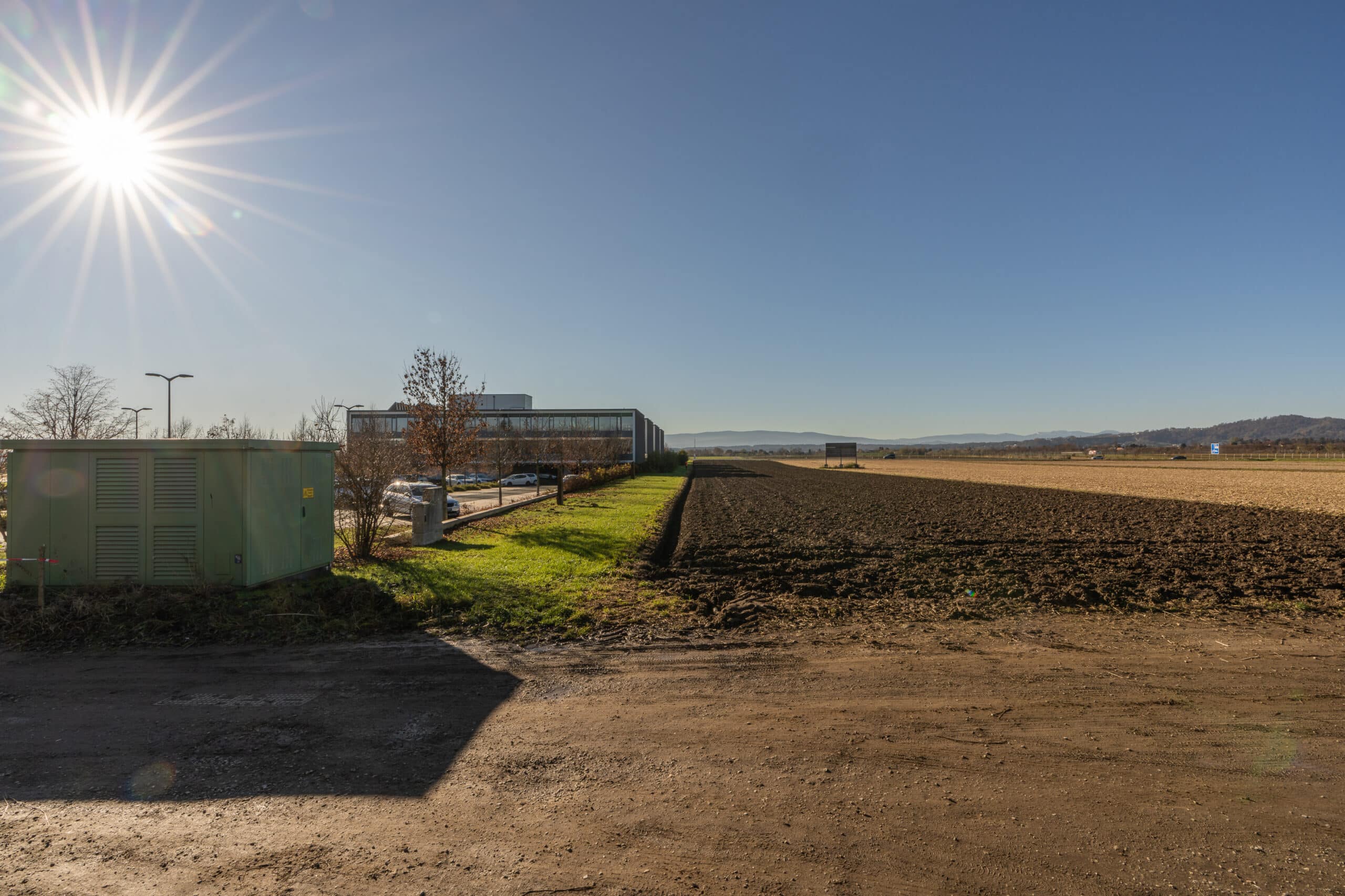Industriegelände bei Sonnenschein mit angrenzendem Feld und Gebäuden am Horizont.