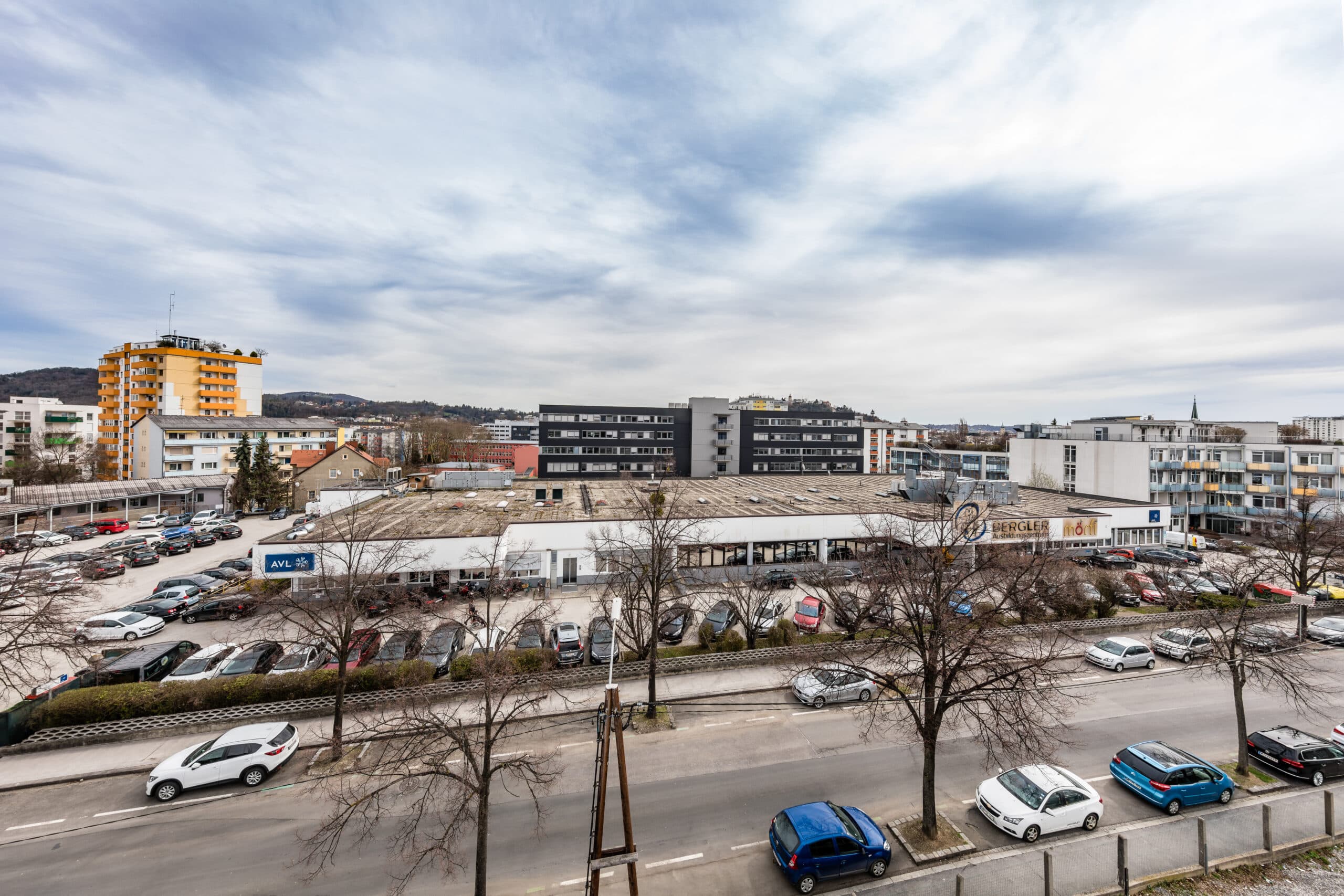 Straßenszene mit geparkten Autos vor Geschäfts- und Wohngebäuden, große freie Parkflächen im Vordergrund.