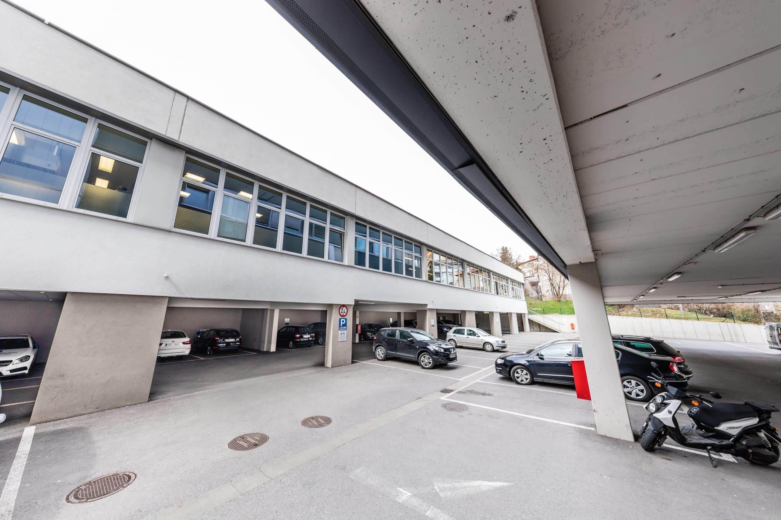 Parkplatz unter modernem Gebäude mit großen Fenstern und parkenden Autos, helle Fassade.