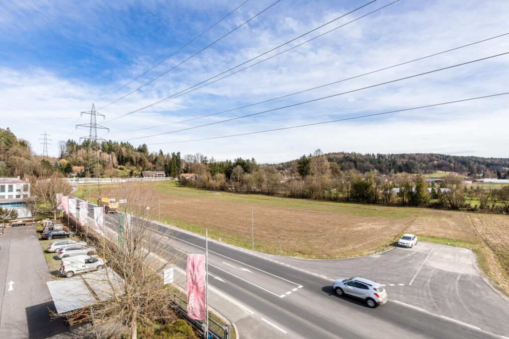 Weitläufige Landschaft mit Feldern und Straßen nahe Grambach, bewölkter Himmel, Stromleitungen sichtbar.