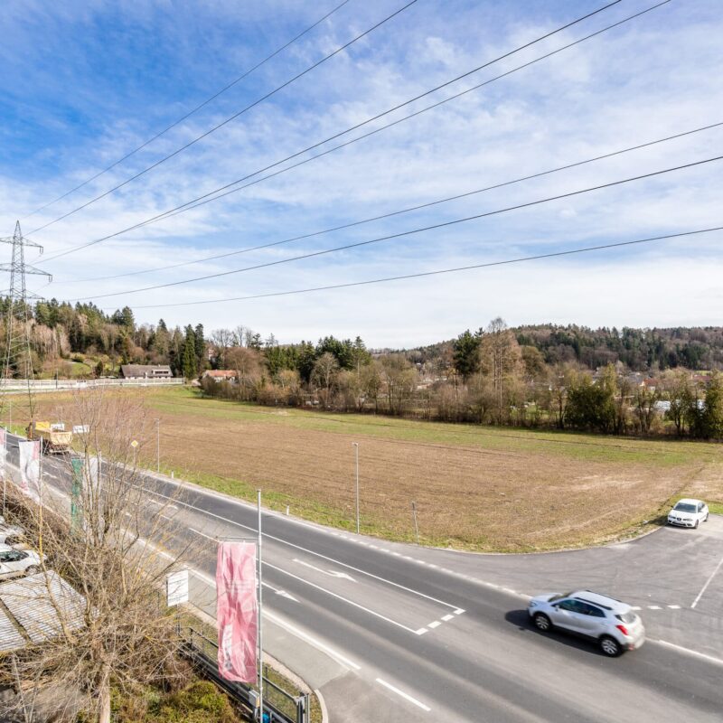 Weitläufige Landschaft mit Feldern und Straßen nahe Grambach, bewölkter Himmel, Stromleitungen sichtbar.