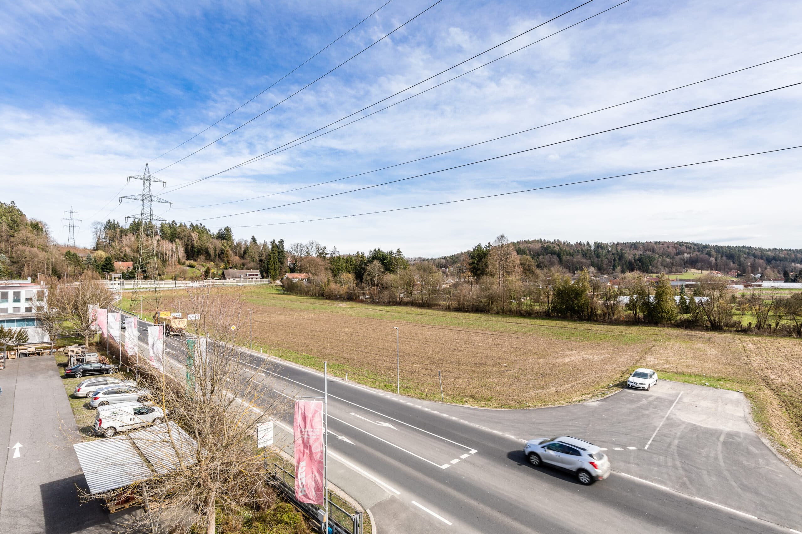 Weitläufige Landschaft mit Feldern und Straßen nahe Grambach, bewölkter Himmel, Stromleitungen sichtbar.