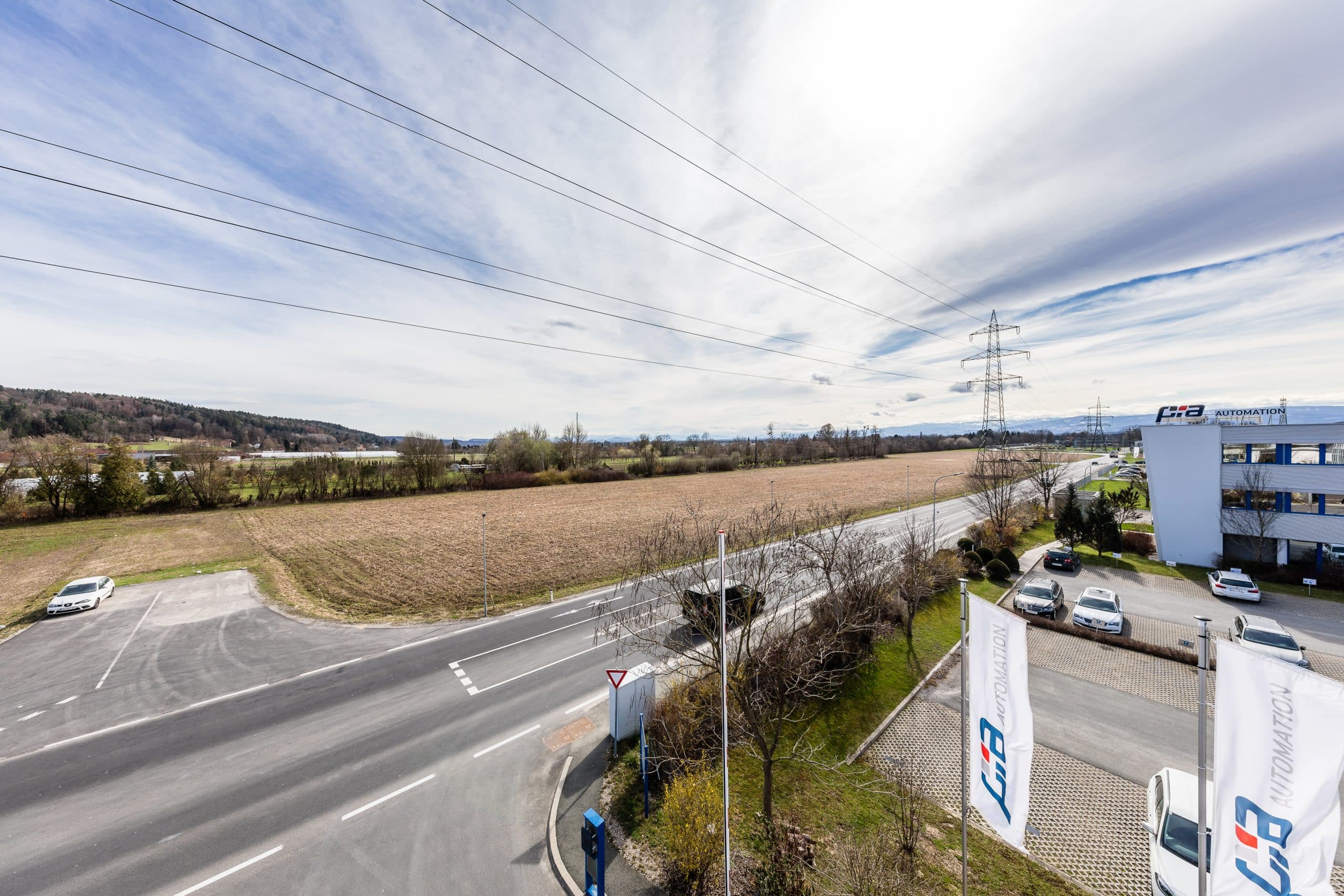 Landstraße bei Grambach mit Feldern und einem modernen Gebäude im Hintergrund, umgeben von Stromleitungen.