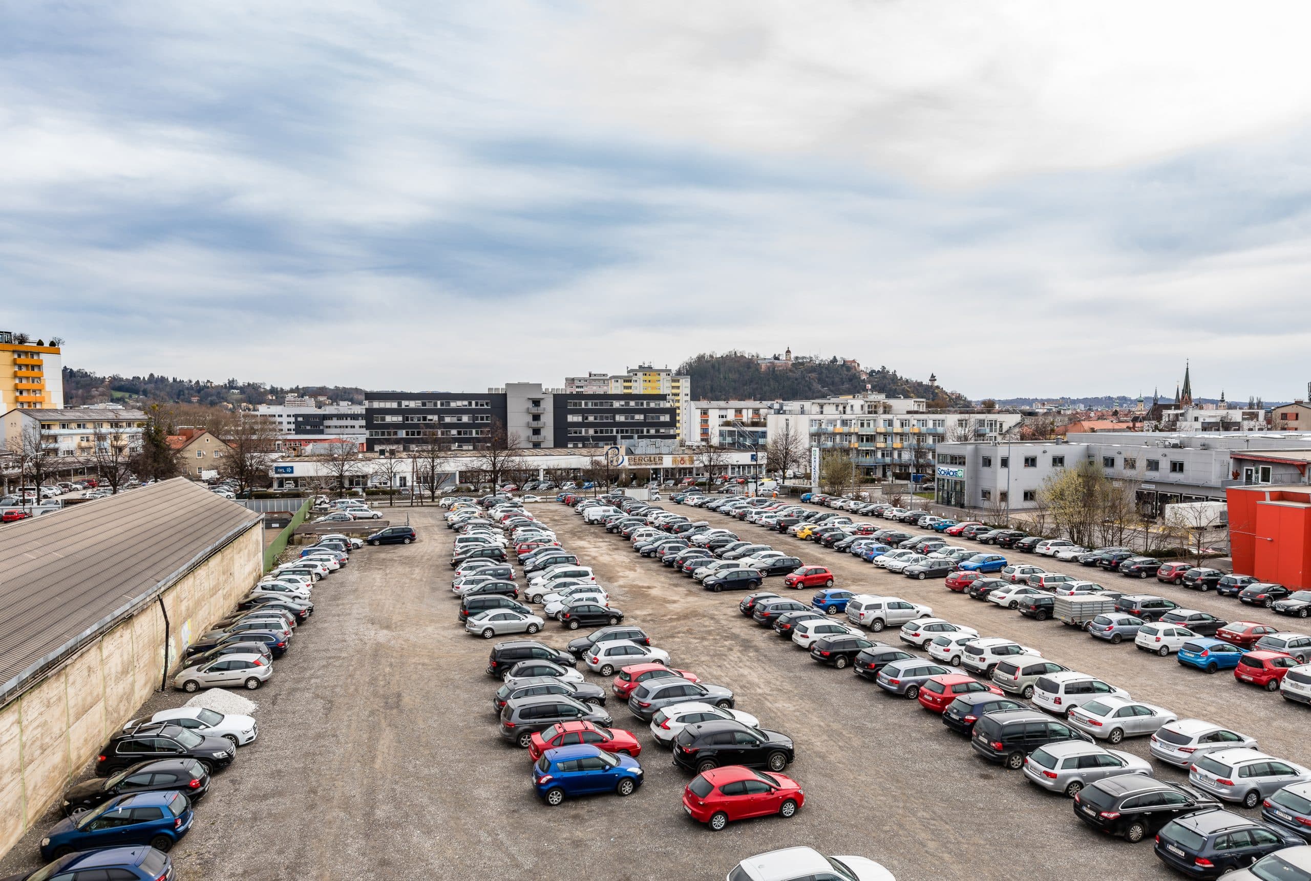 Großer Parkplatz mit vielen Autos, städtische Umgebung und bewölkter Himmel, Gebäude im Hintergrund.