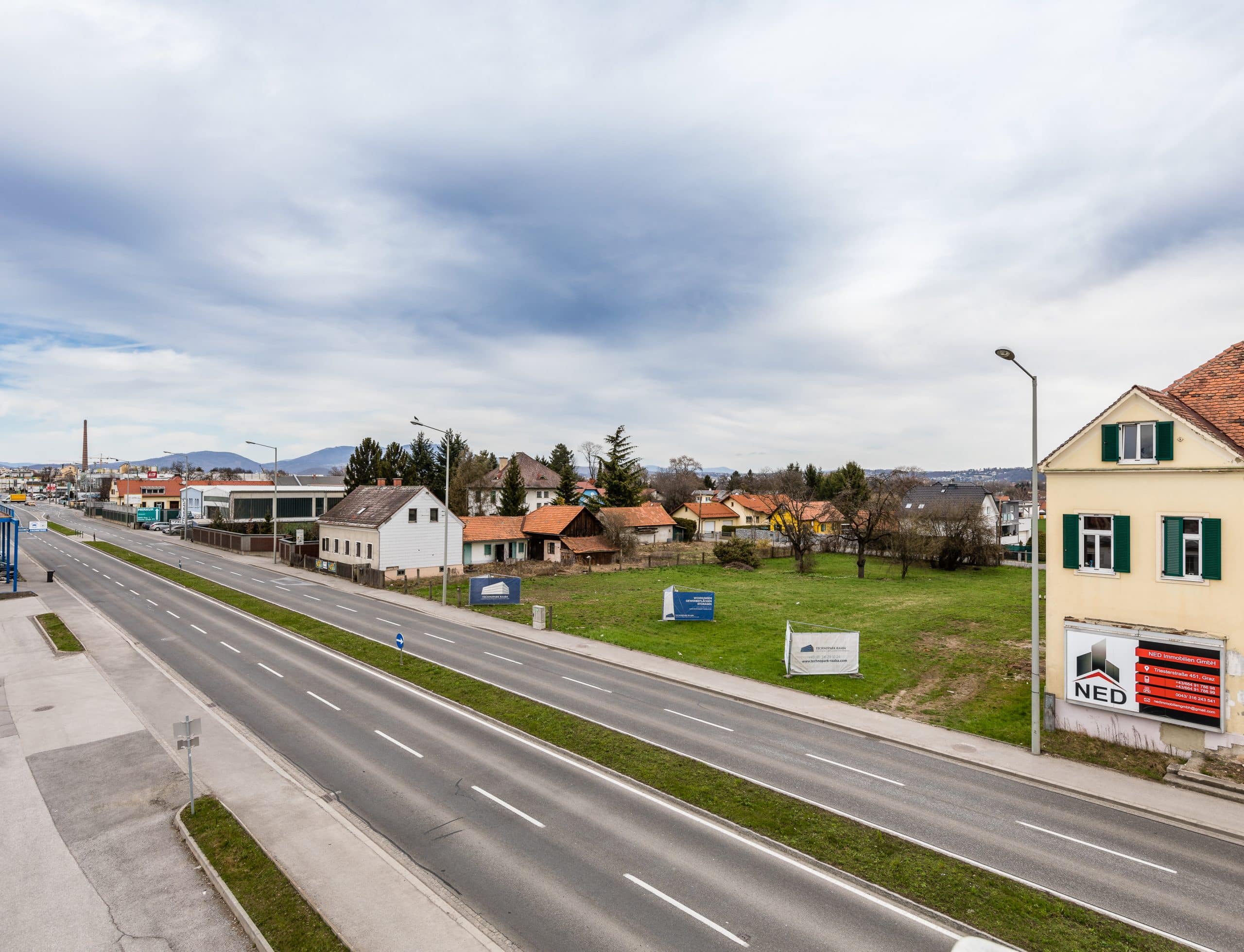 Breite Straße neben Wohn- und Gewerbegebäuden, grüne Wiese im Vordergrund, leicht bewölkter Himmel.