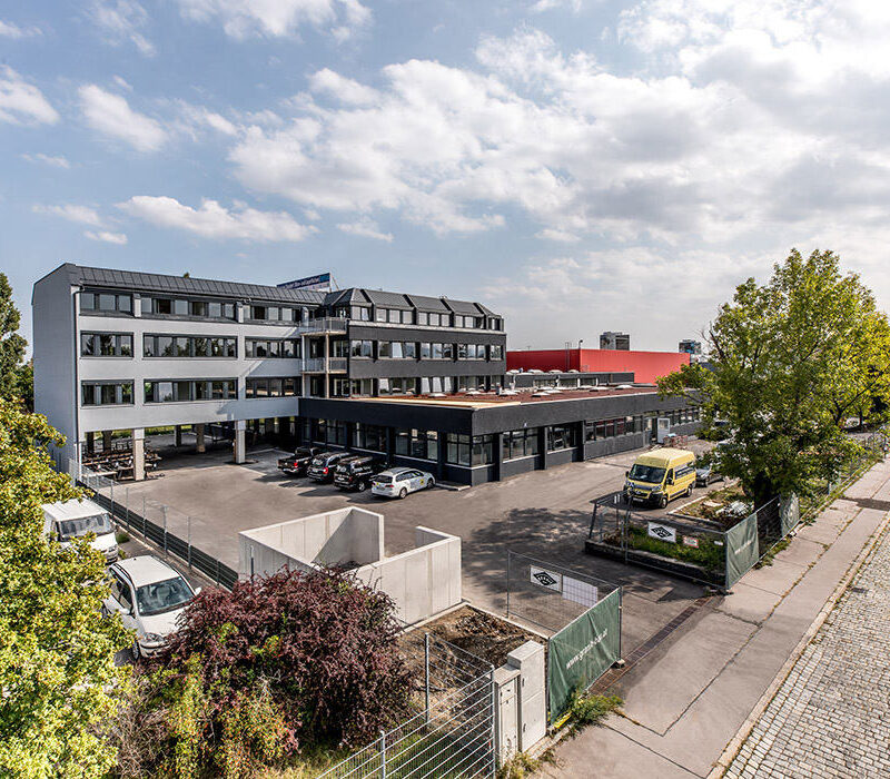 Bürogebäude mit angrenzendem Parkplatz und Bäumen in einer urbanen Umgebung bei sonnigem Wetter.