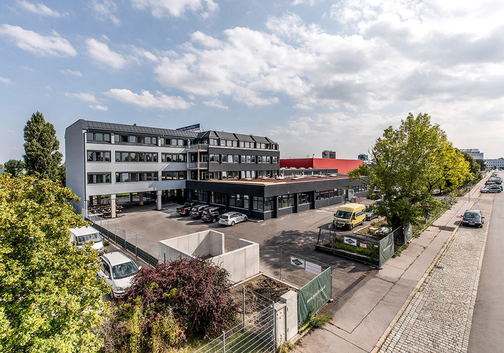 Bürogebäude mit angrenzendem Parkplatz und Bäumen in einer urbanen Umgebung bei sonnigem Wetter.