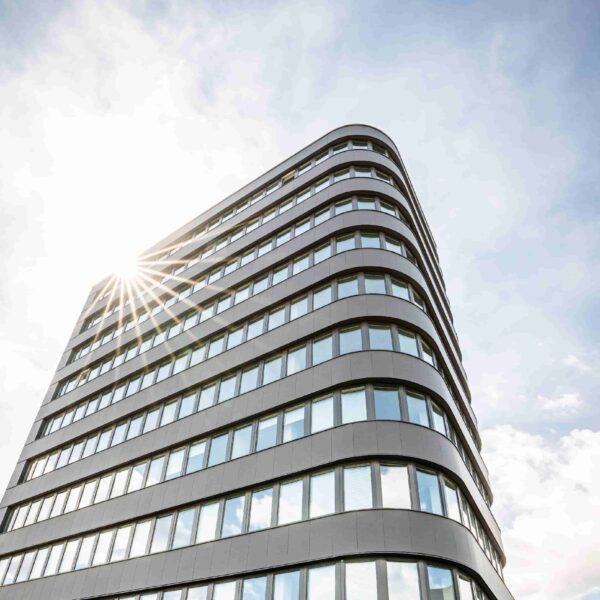 Moderne, mehrstöckige Bürogebäude mit Glasfassade und Sonnenstrahlen am Himmel.