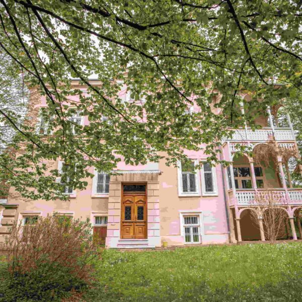 Große Altbauvilla mit Holzpergola, rosa Fassade und grüner Gartenanlage im Vordergrund.