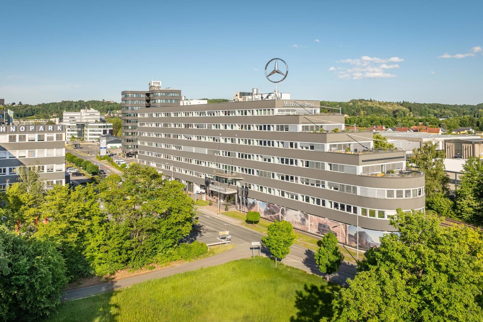 Technopark Raaba TP 2, modernes Bürogebäude mit Mercedes-Logo, umgeben von Grünflächen bei klarem Himmel.
