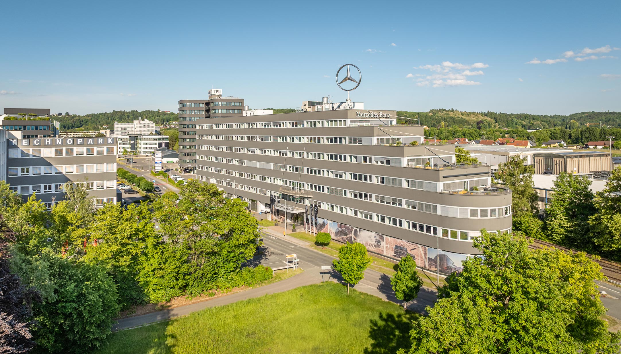 Technopark Raaba TP 2, modernes Bürogebäude mit Mercedes-Logo, umgeben von Grünflächen bei klarem Himmel.