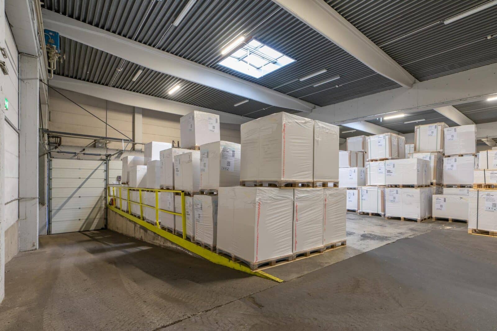 Lagerhalle mit gestapelten Paletten und Kartons auf Rampe in industrieller Umgebung.