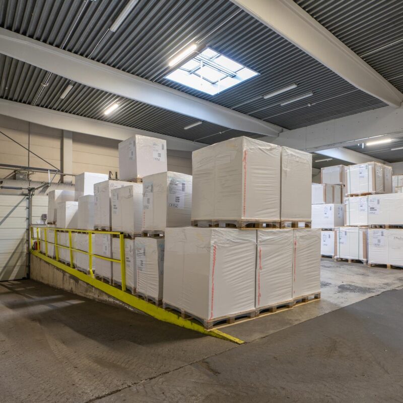 Lagerhalle mit gestapelten Paletten und Kartons auf Rampe in industrieller Umgebung.